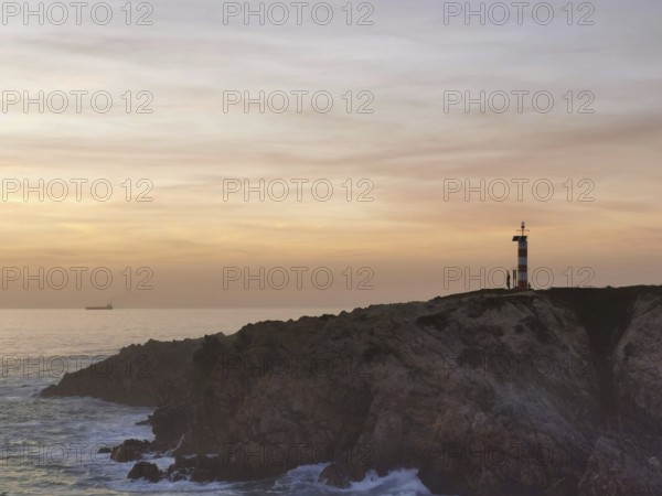 Landscape with lighthouse on rocks, illuminated by sunset, with calm sea and clouds in the background