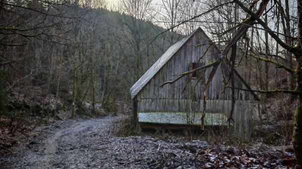 Snowy trail leads to a secluded wooden hut in the winter forest in front of a range of hills, mystical, romantic, Franconian Forest