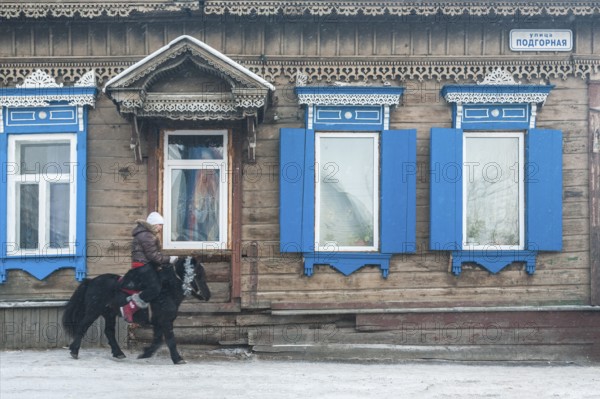 Irkutsk, Siberia, Russia. A young woman riding a pony in front of traditional wooden houses during snow