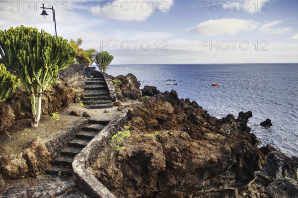 A rocky coastal path with a staircase leads to the sea under a cloudy sky