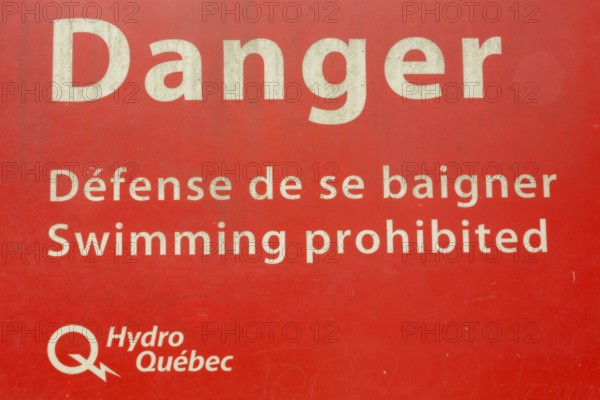 Bilingual French and English Danger Swimming Prohibited in white letters on red painted metal public notice sign posted by Hydro-Quebec utility in public park, Quebec, Canada