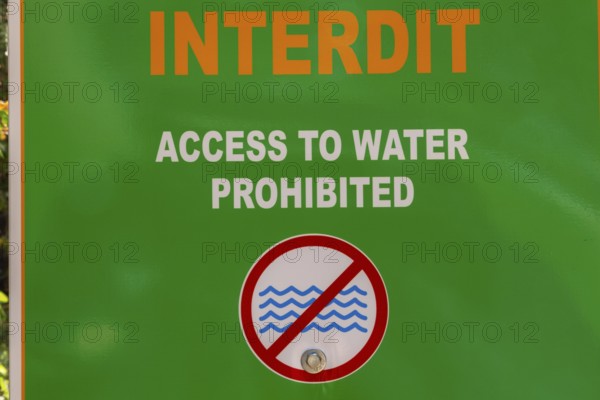 Posted bilingual French and English language warning Access to Water Prohibited in white and orange lettering plus water symbols in red circle on green painted metal pictogram notice sign, Quebec, Canada
