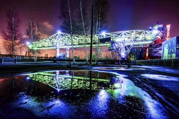 Intensely illuminated bridge construction at night with reflection in water and dramatic clouds, Landschaftspark Duisburg Nord, Duisburg, North Rhine-Westphalia, Germany