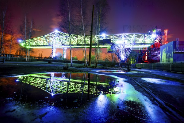Illuminated bridge at night with blue and green lighting reflecting in a pool of water, Landschaftspark Duisburg Nord, Duisburg, North Rhine-Westphalia, Germany