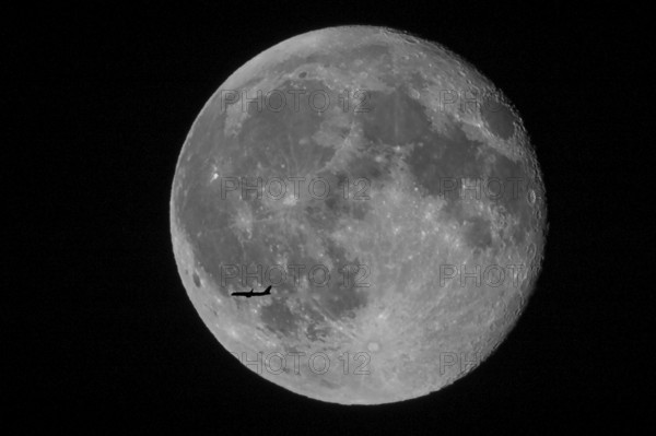 Silhouette of a passenger plane flying in front of a huge and brightly lit full moon with discernible lunar craters in the night sky, North Rhine-Westphalia, Germany