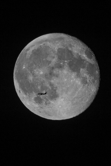 Silhouette of a passenger plane flying in front of a huge and brightly lit full moon with recognizable lunar craters in the night sky, North Rhine-Westphalia, Germany