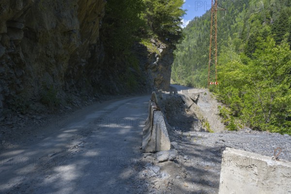 A narrow mountain road leads between rocky cliffs and green forest. Sun and shade areas alternate, destroyed road 7 between Khaishi and Tsalanari, Mingrelia region and Upper Svaneti, Georgia