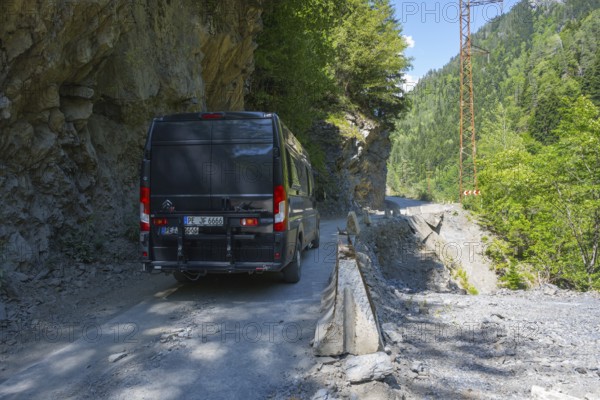 A black van drives on a narrow mountain road surrounded by rocks and thick vegetation, camper drives on destroyed road 7 between Khaishi and Tsalanari, Mingrelia region and Upper Svaneti, Georgia
