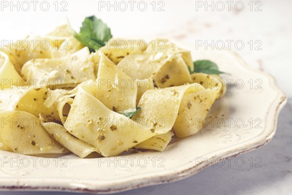 Fresh papardelle pasta, with basil and pesto sauce, is served, on a white plate, with a fork held by hand