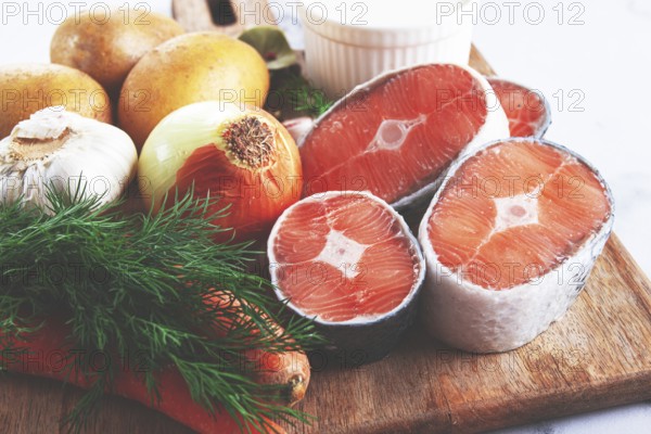Potatoes, salmon, garlic, onion, and herbs are placed on a wooden cutting board. These ingredients are ready for meal preparation in a kitchen