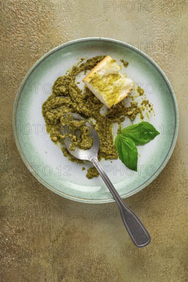 On a platter is a pesto sauce made from fresh basil, with a slice of bread and a spoon on a plate. The pesto sauce looks creamy and green, and the bread is partially eaten
