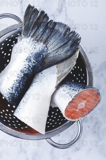 Fish tails are placed in a colander on a marble counter. The colander allows water to drain away from the fish. The fish looks fresh and clean, ready for cooking or preparation