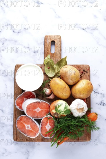 Potatoes, salmon, garlic, onion, and herbs are placed on a wooden cutting board. These ingredients are ready for meal preparation in a kitchen