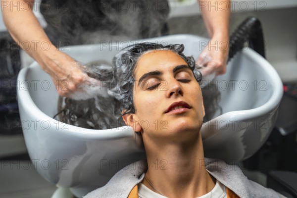 Young woman relaxing with closed eyes while hairdresser hands gently wash her hair with shampoo and warm water, creating steam in a professional salon environment