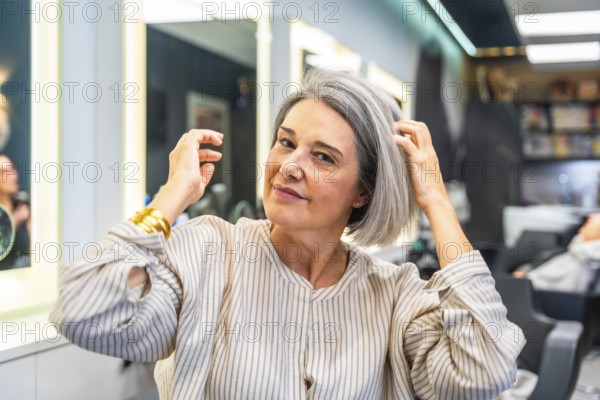 Senior woman smiling and touching her healthy gray hair, sitting in a modern hair salon, ready for a new hairstyle, embracing natural beauty and self care