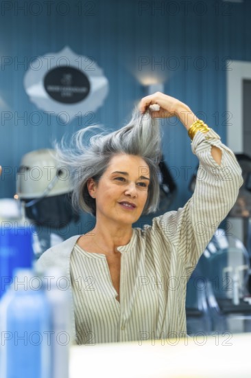 Mature woman looking at her reflection and touching her natural gray hair after a recent blow dry at a modern hair salon, feeling confident and beautiful
