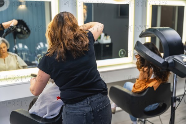 Hairdresser working on a client's hair in a professional beauty salon, with another client undergoing hair treatment in the background, showing a daily scene of hair care services