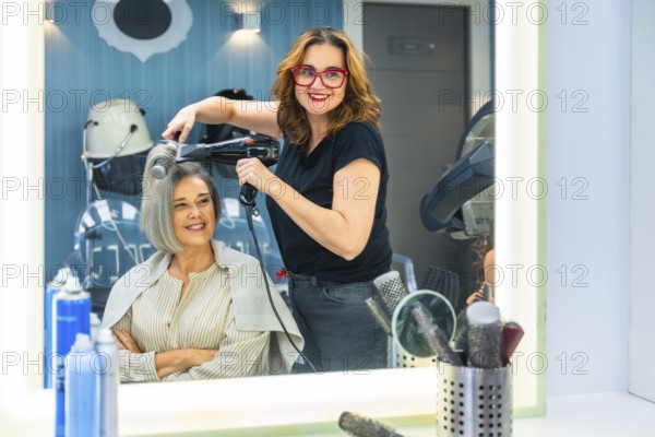Female hairdresser smiling at camera while blow drying a mature customer's hair in a modern salon, creating a stylish look with professional tools and attentive service