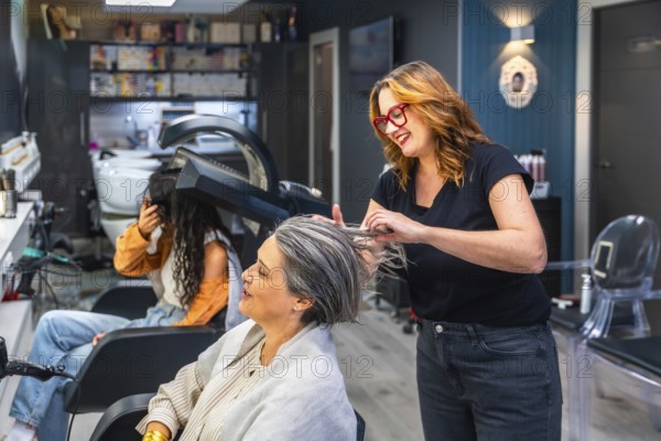 Hairstylist smiling while prepping a senior woman's hair for treatment as another client dries under a hooded dryer in a bright, modern salon setting, relaxed and cheerful