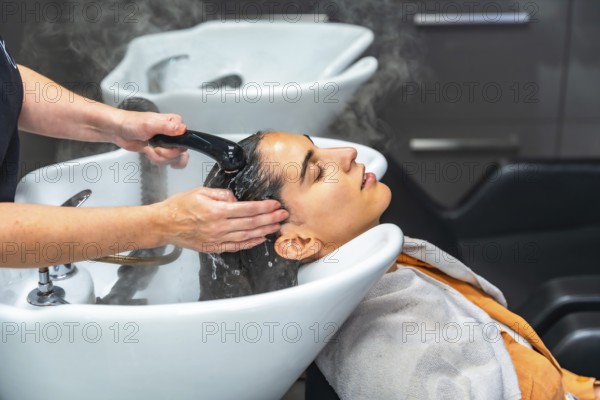 Woman relaxed in salon basin as hairstylist rinses shampoo and massages scalp with warm water and foam, creating a tranquil, pampering hair treatment moment