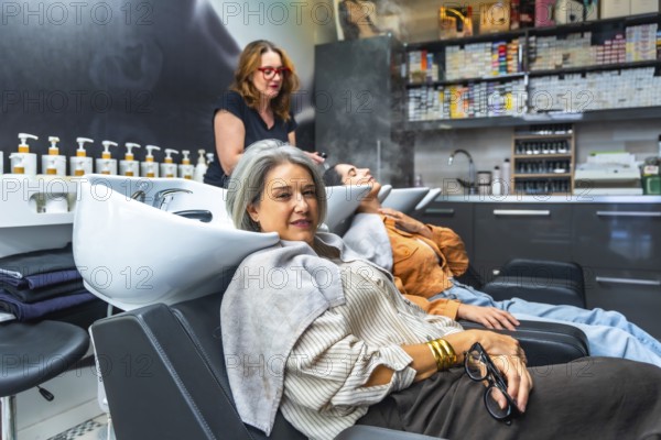 Two women reclining at salon basins while a stylist performs professional shampoo and scalp care, conveying relaxation, pampering, and modern haircare for diverse, mature clients