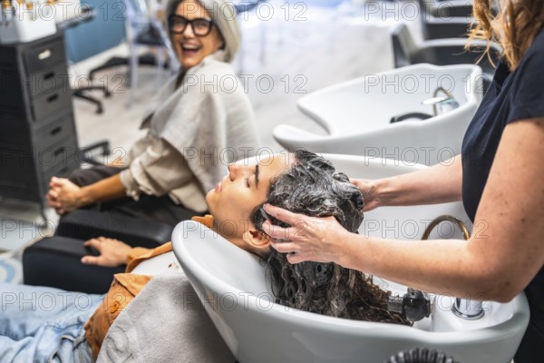 Hairdresser washing customer's hair at basin, providing relaxing care while another woman smiles happily in the background, a common scene in a modern beauty salon