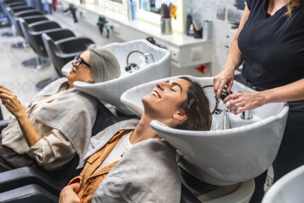 Female hairdresser washing a young woman's hair in a salon hair wash basin, while another client waits nearby, enjoying a relaxing moment during a beauty treatment
