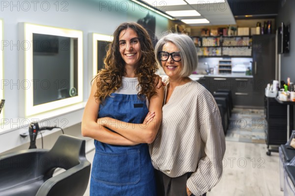 Two cheerful women hairstylists, one younger in an apron and one older with gray hair, standing side by side in a modern salon, smiling confidently as business partners