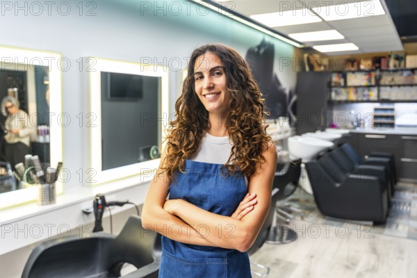Confident young woman with curly hair wearing an apron and smiling, standing with crossed arms in her modern hair salon, representing a small business owner