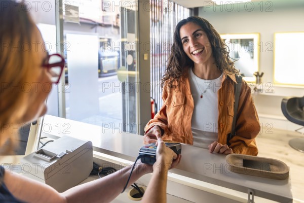 Young woman customer smiles confidently as she makes a secure contactless credit card payment at a modern hair salon reception counter, friendly service interaction
