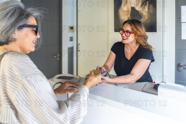 Senior customer making a contactless credit card payment at the reception desk to a smiling female stylist after receiving a beauty service in a modern hair salon