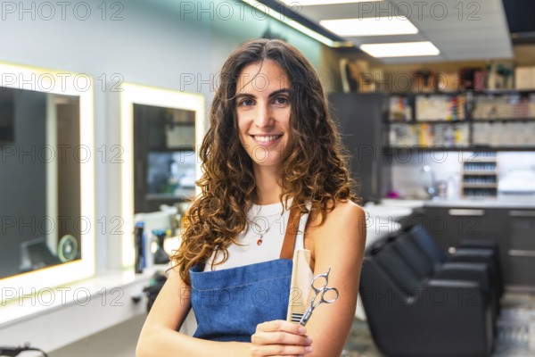 Professional woman with curly hair wearing an apron and holding hairdressing scissors and a comb, standing inside a modern hair salon and smiling confidently