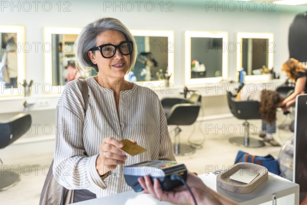 Smiling mature woman paying for hair services by tapping a credit card on a contactless payment terminal at a modern hair salon, completing her beauty treatment