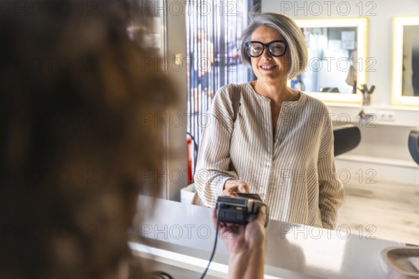 Senior woman smiling while making a digital payment with a credit card at a salon's reception desk, embracing modern transaction technology for services