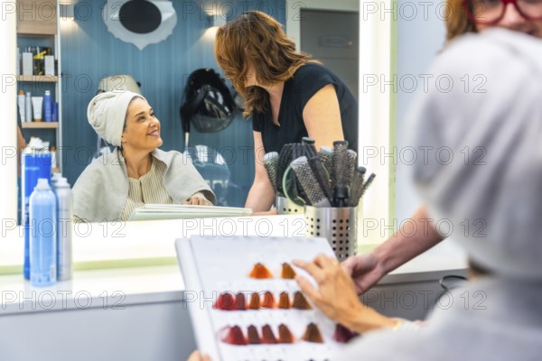 Woman client in a salon chair with a towel wrapped around her hair, looking at her reflection and smiling while a hairdresser helps her choose a new hair dye color from a swatch book