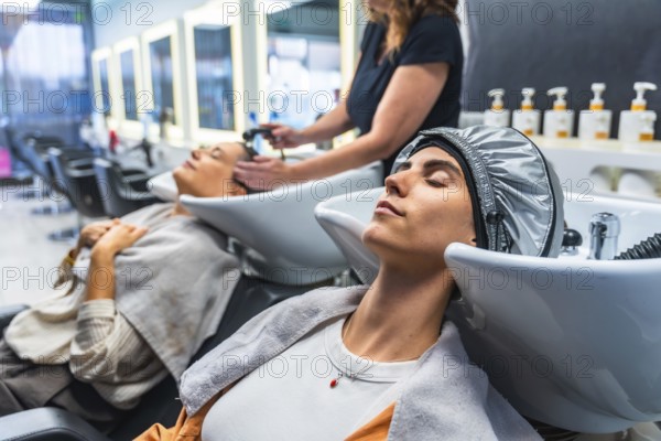 Woman relaxing with eyes closed while receiving a hair treatment and shampoo service at a beauty salon, with another client in the background getting hair washed