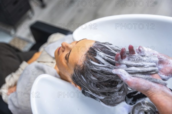 Mature woman relaxing with eyes closed as a hairdresser shampoos her long dark hair over a salon wash basin, creating rich suds and a soothing head massage