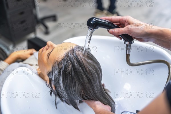 Woman relaxing as hairdresser rinses her hair over a salon sink with a handheld shower, receiving professional shampoo treatment and pampering care for clean, healthy hair