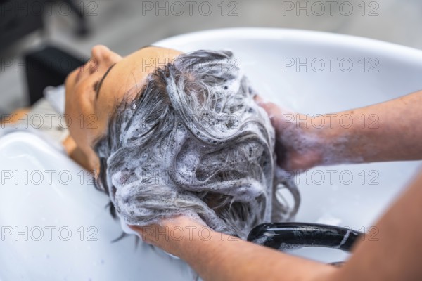 Professional hairdresser washing female client's long dark hair with shampoo and rich foam in a white salon basin, offering relaxing, hygienic beauty care and pampering service