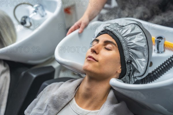 Woman lying at a hair washing sink, receiving a moisturizing hair steaming treatment from a professional hairdresser in a beauty salon, symbolizing care, relaxation, and wellness