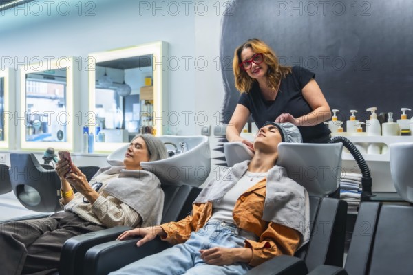 Hairdresser washing young woman's hair at a sleek salon basin while another client relaxes on her phone, capturing a modern, professional pampering and wellness moment