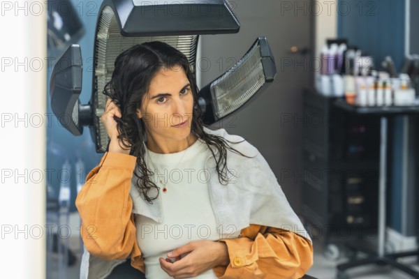 Young woman seated under a professional salon hair dryer, receiving a pampering hair treatment while relaxing, showcasing modern styling, confidence and clean, healthy hair results