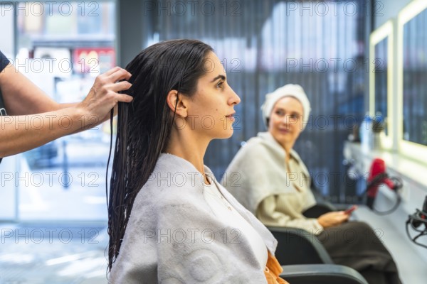 Hairdresser applying hair treatment to a young woman's wet, dark hair, highlighting beauty, care, and wellness services at a modern salon, with another client waiting in the background