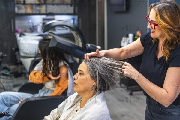 Hair stylist applying product to a senior woman's hair in a professional salon setting, with another customer under a hair dryer in the background, focusing on beauty care and pampering