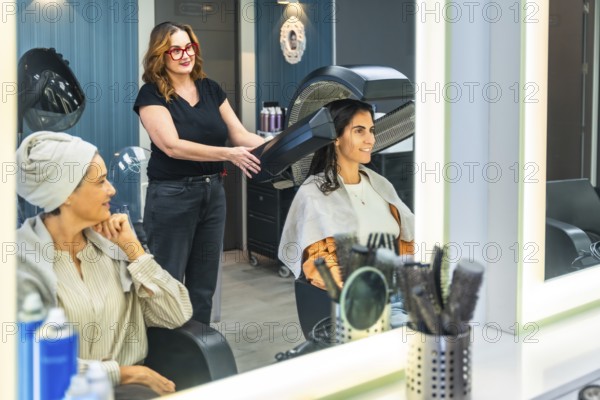 Hairdresser working on a client's hair with a professional bonnet dryer in a modern salon while another woman waits, reflecting a moment of self care and beauty treatment