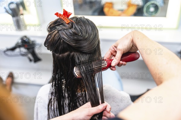 Professional hairdresser brushing through wet, dark long hair with clips, preparing sections for styling in a modern salon, close up of hands, comb, and client care
