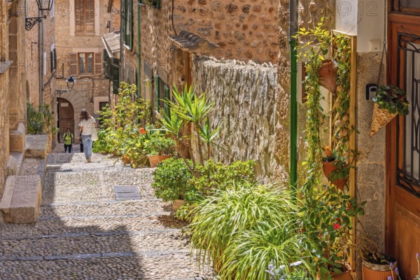 Idyllic cobblestone alley with green plants and flowers at the house walls and walking people a sunny summer day, Fornalutx, Mallorca, Spain