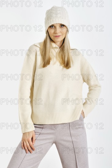 A model stands confidently in a studio while wearing a light sweater and a knitted hat. The setting features bright lighting that highlights her outfit and expression as she poses for the camera