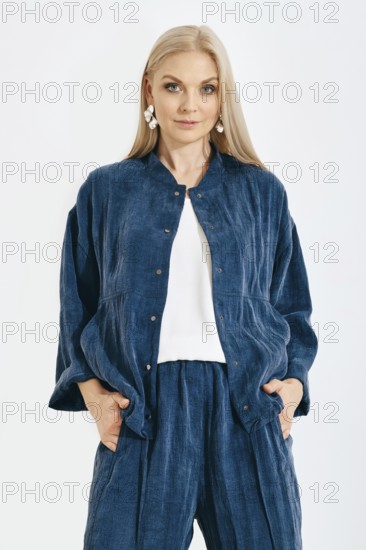 Blonde woman stands with her hands in pockets wearing a denim jacket and matching pants in a studio. She has long hair and is styled simply