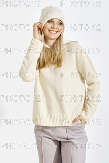 A woman stands smiling while wearing a light sweater and a warm hat. She has long blonde hair and is posing in front of a plain background. The setting looks bright and simple